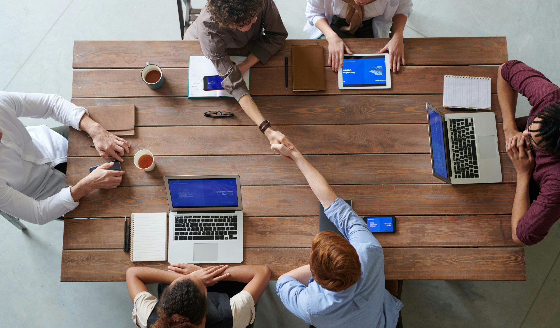 Ein Konferenztisch von oben gesehen, an dem 6 Personen mit ihren Computern sitzen. Zwei von ihnen schütteln sich die Hände.