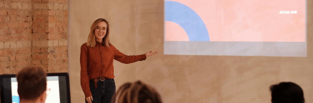 Eine junge Frau hält einen Vortrag vor einem kleinen Publikum und zeigt dabei auf den Bildschirm.