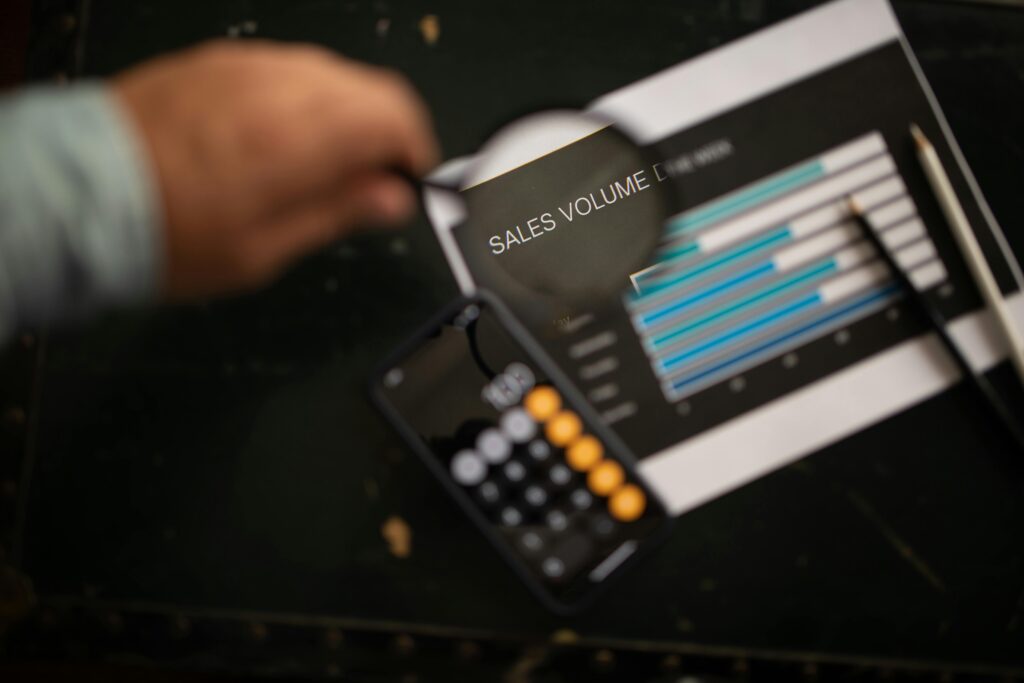 Lupe vergrößert die Überschrift „Sales Volume“ auf einem Balkendiagramm; daneben Taschenrechner und Stift, Hintergrund unscharf.
